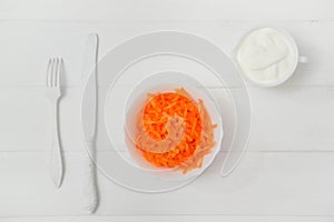 Flat lay, composition, freshly grated carrot in a plate. On the wooden table, a knife and fork. White background. Light