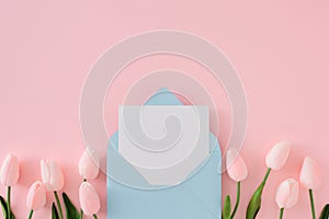 Flat lay composition made of open envelope with white card and spring flowers on pastel pink background.