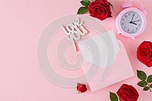 Flat lay composition made of red roses, open envelope with card, inscription love you, alarm clock