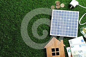 Flat lay composition with solar panel, house model and money on grass. Space for text