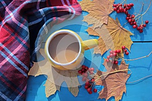 Flat lay composition with yellow cup of coffee, plaid, red berries, and autumn leaves on blue wooden background. Cozy autumn mood.