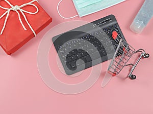 Flat lay of computer keyboard , shopping cart,red gift box , medical mask and alcohol sanitizer gel on pink background with copy