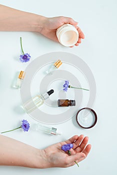 Flat lay of cosmetical flasks and jars betwen two hands with jar of cream and blue flower. Aromatherapy and skin treatment concept