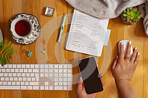 Flat lay of cozy workplace. Schedule planner, keyboard, phone and tea. Female hand holding mouse and phone