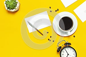 Flat lay cup black coffee, coffee beans, black alarm clock, pens, white cards, succulent on yellow background top view copy space