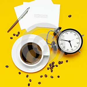 Flat lay cup black coffee, coffee beans, black alarm clock, pens, white cards on yellow background top view copy space. Creative
