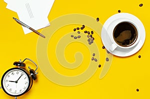 Flat lay cup black coffee, coffee beans, black alarm clock, pens, white cards on yellow background top view copy space. Creative