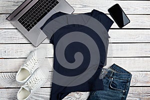 Flat lay of dark blue t-shirt lying on rough wooden planks with laptop, smartphone and apparel accessories.