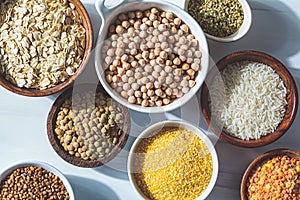 Flat lay of dry cereals, grains and legumes in white and wooden bowls, white background. Vegan protein concept