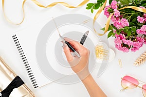 Flat lay fashion office desk. Female workspace with blank paper notebook and hand of woman holding pen, pink rose flowers, golden