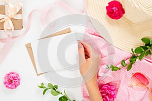 Flat lay fashion office table desk. Female workspace with human hand holding blank paper card, straw hat, rose flower buds, gift