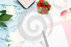 Flat lay feminine home office desk with white peony flowers, strawberry, sunglasses, purse, paper notepad. Top view woman