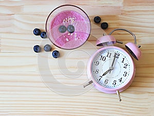 Flat lay of a glass blueberry smoothie and pink vintage alarm clock 7 o`clock on wooden table background