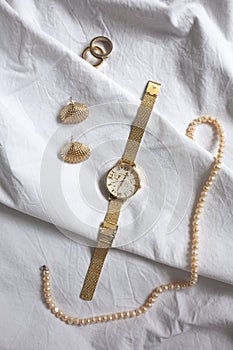 Flat lay gold jewelry on white cotton background: pearl necklace, ring, shell-form earring, watch