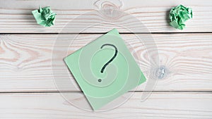 Flat lay of green sticker sticky note with question mark drawn with black marker near crumpled notes on wooden table.