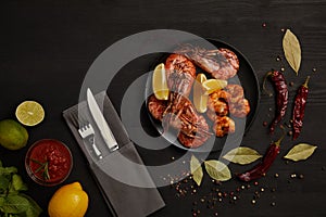flat lay with grilled shrimps with lemon pieces on plate and arranged sauce, spices, ingredients and cutlery around on black surfa