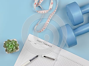 Flat lay of habit tracker book, dumbbells, measure tape and succulent plant pot on blue background with copy space. lose weight,