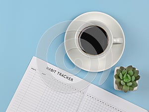 Flat lay of habit tracker book with pen,  cup of black coffee and succulent plant pot on blue background with copy space
