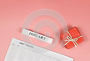 Flat lay of habit tracker book,  red gift box and wooden calendar January  on pink background