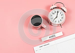 Flat lay of habit tracker book, wooden calendar April, pink  vintage alarm clock 7 o`clock and pink cup of black coffee  on blue