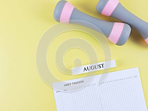 flat lay of habit tracker book, wooden calendar August, gray pink dumbbells, on yellow background with copy space. Self