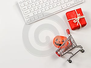 Flat lay of  Halloween pumpkin on shopping cart, red gift box and computer keyboard  on white background with copy space.