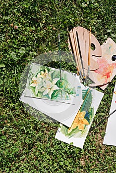 Flat lay of handmade oil paintings of lilies, brushes and palettes on green grass