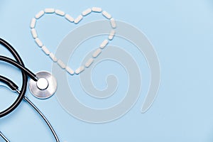 Flat lay. A heart shape made from white medical pills near a doctor's stethoscope on isolated blue background. Ad