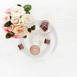 Flat lay home office desk. Female workspace with pink and beige roses flowers bouquet, coffee and chocolates on white