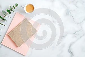 Flat lay home office desk. Female workspace with stylish pink notepad, cup of coffee, eucalyptus leaf on marble table. Top view