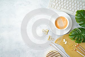 Flat lay home office feminine table with coffee cup, golden accessories, computer keyboard and tropical leaves on grey background