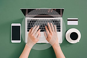 Flat lay of laptop, coffee and notebook on green background. Female hands typing on keyboard