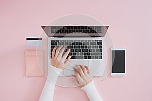 Flat lay of laptop, credit card, mobile phone and passport on pink background. Female hands typing on keyboard