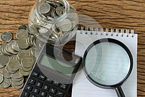 Flat lay of magnifying glass on white note book paper and calculator with stack of coins on wood table as financial saving or tax