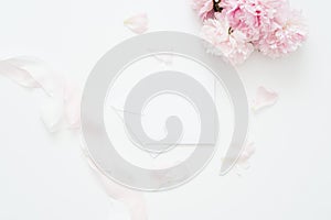 Flat lay, Minimal woman`s desktop with blank page mock up, envelope, peony flower with petals, pale pink silk ribbon.