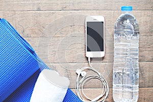 Flat lay of mobile phone with earphones, water bottle, towel and blue yoga mat