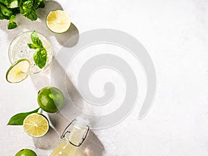 Flat lay of mojito cocktail with lime, fresh mint and ice, bottle with mojito on white texture background close up. Summer