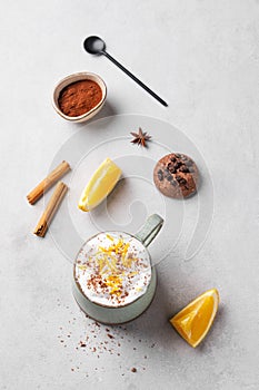 Flat lay of a mug of hot chocolate with whipped cream and orange zest on a light background with a cinnamon sticks, cookies, spoon
