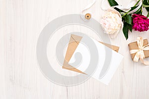 Flat lay peonies flowers, craft envelope, blank paper card, gift box, ribbon on wooden background. Top view feminine workspace,