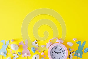 Flat lay photo of blue, white easter eggs, cute rabbits, pink alarm clock and paper bunny toppers on yellow background
