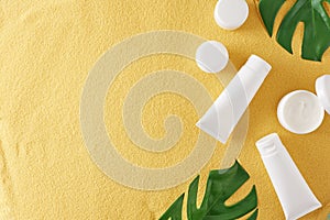 Flat lay photo of cosmetic bottles without label, cream jars and green tropical leaves on sandy background