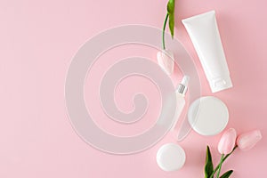 Flat lay photo of cosmetic tubes without label, cream jars, dropper bottle and spring flowers on pastel pink background