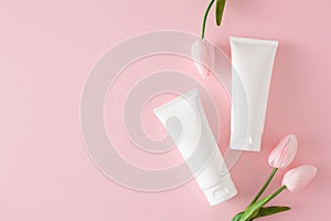 Flat lay photo of cosmetic tubes without label and spring flowers on pastel pink background