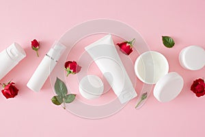 Flat lay photo of cream jars, white tubes without label, red roses and leaves on pastel pink background.