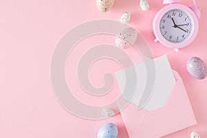 Flat lay photo of Easter eggs, open envelope with white card and alarm clock on pastel pink background