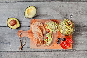 Flat lay photo, freshly prepared guacamole in small glass bowl, bread, tomatoes, olives at working board and two avocados next