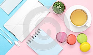 Flat lay photo of office desk with case for phone and tablet, notebook, tea mug, macaroon, cactus, pink and blue background