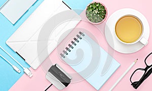 Flat lay photo of office desk with case for phone and tablet, notebook, tea mug, pencil, cactus, pink and blue background