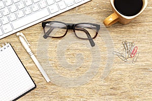 Flat lay photo of office desk with keyboard, notebook, eyeglasses and coffee cup, paper clip and pen.