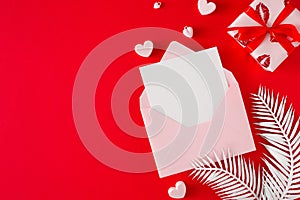 Flat lay photo of open envelope with card, white tropical leaves, gift box and hearts on red background